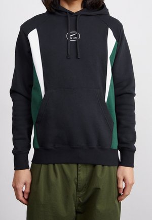 Osoba ubrana w czarną bluzę z kapturem z białymi i ciemnozielonymi bocznymi panelami oraz zielone spodnie, stojąca na jednolitym tle.