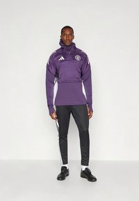 Lilla treningsjakke med halv glidelås, hvite detaljer og logo; kombinert med svarte treningsbukser og svarte sko; sporty design.