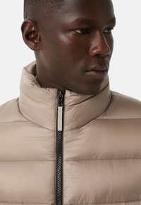 Beige polstret jakke med høj krave, lynlåsdetalje og kontrasterende syninger. Materialet ser glat og blankt ud. Logo synligt på lynlåsgrebet.