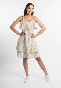 Rochie cu imprimeu floral în crem, cu flori violete, bretele volanate și fustă în straturi. Purtată cu sneakerși albi, evidențiind un stil casual.