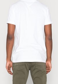 Biała koszulka z krótkim rękawem o prostym zakończeniu, wykonana z bawełny. Połączona z oliwkowo-zielonymi spodniami posiadającymi tylne kieszenie i subtelną teksturę.