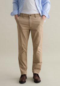 Chino in cotone beige con una vestibilità slim, dotati di tasche laterali, chiusura a singolo bottone e scarpe in pelle marrone. Camicia a righe blu chiaro.