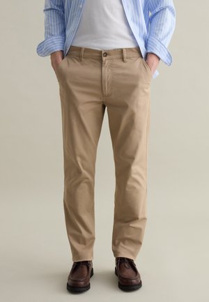 Chino in cotone beige con una vestibilità slim, dotati di tasche laterali, chiusura a singolo bottone e scarpe in pelle marrone. Camicia a righe blu chiaro.