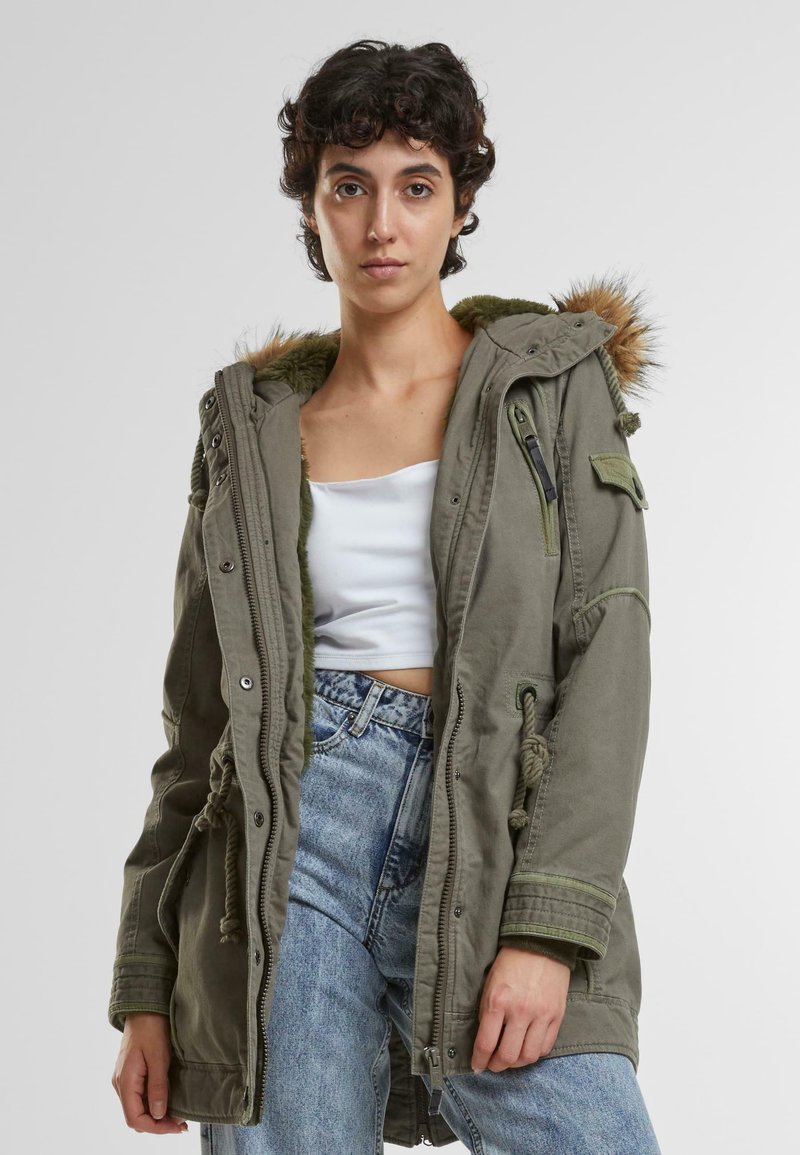 Olijfgroene parka jas met nep-bont afgewerkte capuchon, trekkoord in de taille, meerdere zakken en ritssluitingen. Gedragen over een witte crop top en jeans.