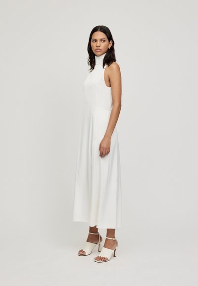 Vestido blanco sin mangas con cuello alto, de corte relajado y tejido fluido. Combinado con tacones abiertos de color crema con tiras en el tobillo y un ligero tacón.