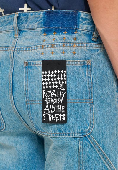Lyseblå denimjeans med orange krydssting-accenter og et sort mærke med hvid tekst, der står "ROYALTY HEROISM AND THE STREETS."