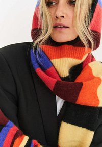 Wełniany szalik z wyrazistymi, wielokolorowymi paskami w kolorach czerwonym, czarnym, pomarańczowym, fioletowym i żółtym; miękka faktura; noszony z dopasowanym kapturem.