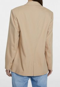 Blazer beige a righe con vestibilità su misura, apertura singola sul retro e spalle strutturate, abbinato a pantaloni in denim azzurro chiaro.
