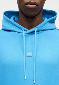 Nærbilde av en person som har på seg en lys blå hettegenser med Calvin Klein-logoen plassert midt under halskanten og med hengende snorer.