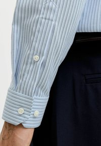 Hele sinine triibuline särkvarrukas valgete nuppudega. Kangal on sile tekstuur ja kitsad vahelduvad tumedad triibud. Mansett on korralikult kokku keeratud.