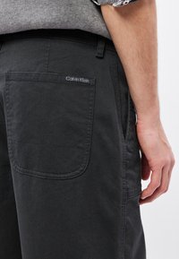 Sötétszürke pamut nadrág hátul varrott logós zsebbel. A dizájn tiszta vonalakat tartalmaz, további minták nélkül.