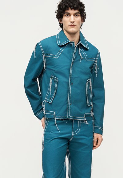 Νέος άνδρας φοράει μπλε-πράσινο τζιν μπουφάν και παντελόνι με αντίθετες λευκές ραφές και φθαρμένες άκρες, στέκεται μπροστά σε απλό φωτεινό φόντο.