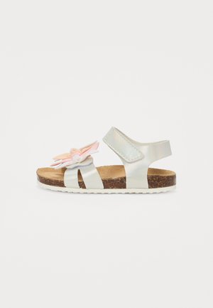 Sandale argentée claire avec une semelle en liège, mettant en avant un accent floral décoratif en rose et blanc doux, une sangle ajustable et un design ouvert.