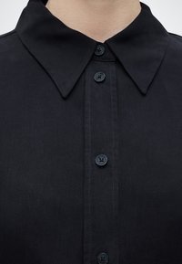 Κοντινή λήψη μαύρου υφασμάτινου γιακά πουκάμισου με τέσσερα ορατά μαύρα κουμπιά κλειστά, εμφανίζοντας την περιοχή του άνω στήθους και του λαιμού.