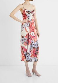 Žena v midi šatech bez rukávů s květinovým potiskem, doplněná o průhledné béžové ponožky a lila sandály na kitten heels.