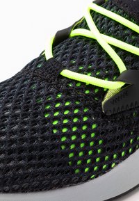 Musta verkkokangassneakersit neonvihreillä nauhoilla ja yksityiskohdilla, teksturoidulla kankaalla, jossa on hengittävä kuvio, ja sileällä valkoisella pohjalla.