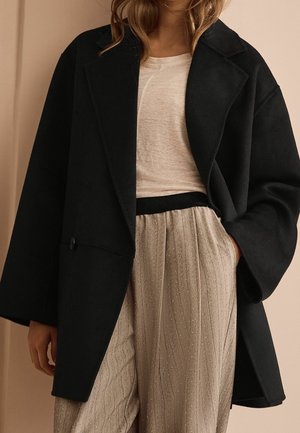 Černý oversize kabát z vlněné směsi s jedním knoflíkem, široké rukávy, nošený přes světle béžový top a texturované, plisované kovové kalhoty.