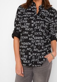 Chemise noire à boutons avec un motif de texte blanc comportant des phrases. Manches courtes retroussées, tissu lisse, col classique et poches à l'avant.