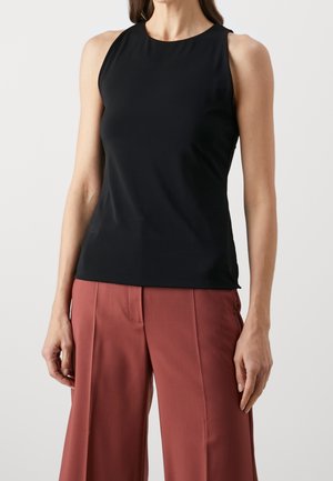 Femme portant un haut noir sans manches à col montant et un pantalon ample couleur rouille, debout devant un fond clair uni.