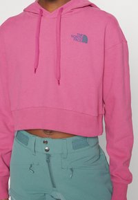 Cappotto rosa accorciato realizzato in tessuto morbido, con cappuccio regolabile e un logo blu sul petto, abbinato a pantaloni verde menta.