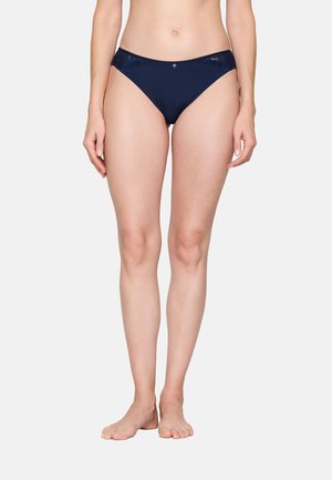 Woman wearing navy blue bikini-style underwear with lace details, standing barefoot against plain light background.