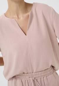 Tuta rosa pallido con scollatura a V, maniche corte e vita elasticizzata. Presenta un tessuto morbido e testurizzato con dettagli di cucitura minimi.