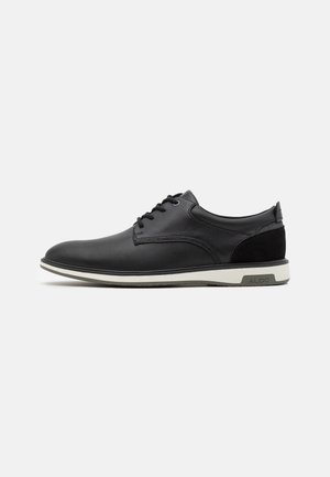 Zapato para hombre negro de caña baja con cordones, paneles laterales texturizados, suela blanca y detalle de marca gris en el talón.