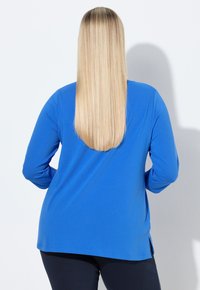 Niebieska bluzka z długim rękawem o luźnym kroju, wykonana z miękkiego materiału. Posiada zaokrąglony dół i rozcięcia po bokach dla większej swobody ruchów.