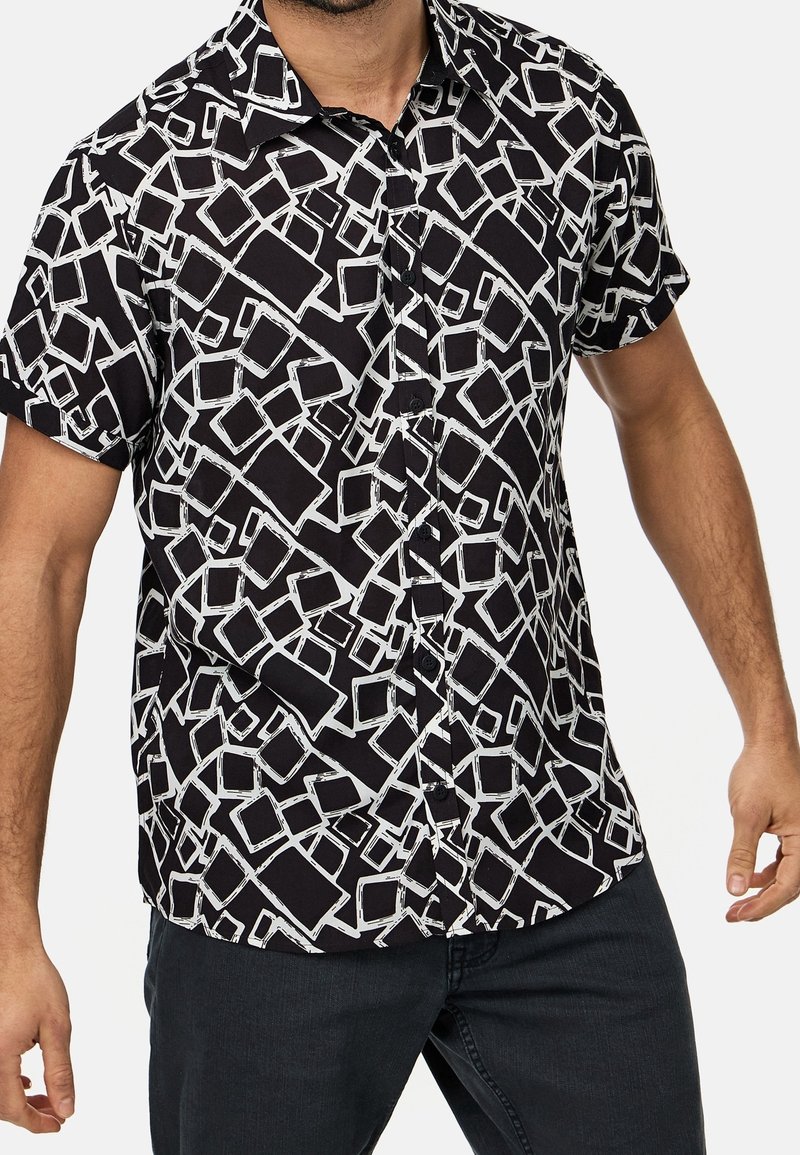 Korte mouwen knoop-overhemd van zwart stof met een wit abstract geometrisch patroon. Beschikt over een kraag en knopen aan de voorkant.