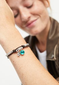 Pulsera de cuero con cuentas de plata, un charm de turquesa translúcido y un colgante de trébol de plata, sobre un tono de piel claro.