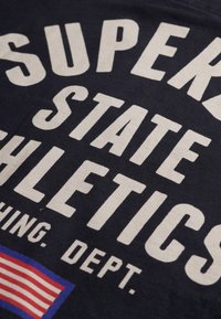 Navyblaues T-Shirt mit auffälligem, cremefarbenem Text "SUPER STATE ATHLETICS" und einem Grafikdesign der amerikanischen Flagge in Rot, Weiß und Blau.