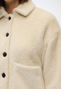 Kreminė dirbtinė kailio striukė su apykakle, su juodais mygtukais ir priekinėmis kišenėmis. Audinys turi švelnų, tekstūruotą paviršių.