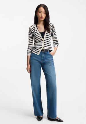 Donna che indossa un cardigan a righe bianche e nere, top nero, jeans blu a vita alta e gamba larga e scarpe nere appuntite, in piedi con una mano in tasca.