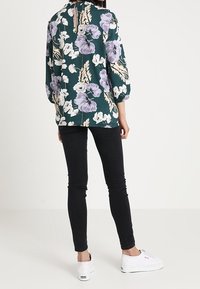 Blomtryckt blus i grönt med lila och vita blommor, med puffärmar och en nyckelhålsgrej på baksidan, tillsammans med svarta skinny jeans och vita sneakers.
