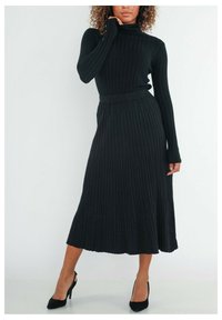 Robe en maille côtelée noire avec col montant, manches longues et taille ceinturée, descendant jusqu'à un ourlet midi, associée à des escarpins noirs.