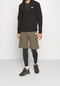 Giacca in pile nera con zip, pantaloni corti oliva con cerniere, leggings neri e scarpe da trekking marroni. Marchi distintivi sulla giacca e sui pantaloni.