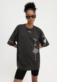 Frau mit lockigem Haar, die schwarze Sonnenbrillen trägt, ein übergroßes schwarzes T-Shirt mit weißen Sonnen- und Skorpionsmotiven und den Worten "MIND OVER MATTER" sowie schwarze Radfahrshorts.