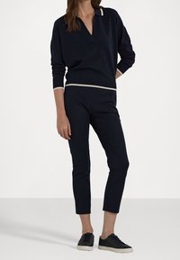 Marinblå stickad tröja med V-ringning och kontrasterande elfenbensfärgade detaljer, ihop med marinblå figurnära byxor och mörka sneakers. Slät textur.