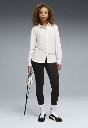 Kobieta stojąca, ubraną w białą bluzkę z paskiem w gwiazdy, czarne legginsy, czarno-białe tenisówki, białe skarpetki i trzymająca małą białą torebkę.