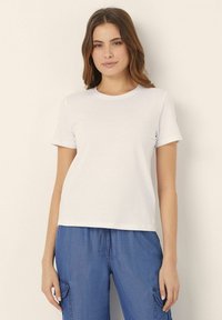 Un tricou alb cu maneci scurte, fabricat din bumbac. Design clasic cu guler rotund, cu o textură netedă, asociat cu pantaloni albaștri cu șnur pentru o ținută lejeră.