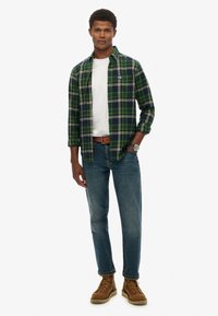 Grün-karierte und marineblaue Flanellhemd über einem weißen T-Shirt, kombiniert mit mittelblauen Jeans und braunen Schnürstiefeln. Strukturierter Stoff, lässige Passform.