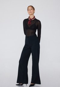 Schwarzes Langarmoberteil mit transparenter Textur und Rundhalsausschnitt, kombiniert mit hochgeschnittenen, schwarzen weitem Hosen. Accessoires: ein kastanienfarbener Schal.