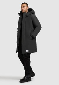 Zwarte, geïsoleerde parka met capuchon, twee zijzakken en een merkpatch op de onderkant. Draagt over zwarte broek en zwarte laarzen.