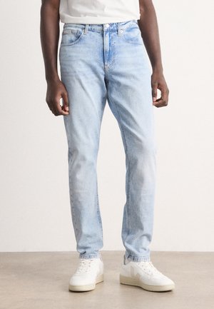 Personne portant un jean bleu clair, des baskets blanches et un t-shirt blanc, debout sur un sol uni face à un mur blanc.