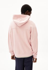 Felpa con cappuccio rosa, dal taglio rilassato, con una texture morbida, cuciture visibili e un orlo inferiore. Indossata sopra pantaloni blu scuro, senza vista frontale.