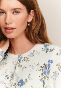 Cardigan floral en tricot blanc doux, ornée de fleurs bleues et vertes. Col arrondi, fermeture à boutons et manches longues avec des poignets côtelés.