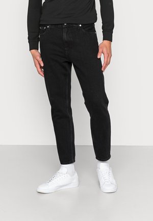 Personne portant un jean slim noir, des chaussettes blanches et des baskets blanches, debout les mains détendues le long du corps devant un fond uni.