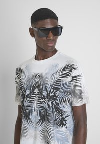 Camiseta blanca con un estampado gráfico de hoja de palma en negro y azul, combinada con gafas de sol negras de forma angular, con un acabado suave y un diseño audaz.