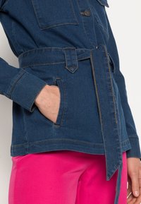 Denimjacka i mörkblått med bälte i midjan. Har dubbla bröstfickor, knappstängning och kontrasterande sömmar.