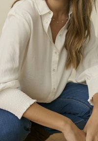 Femme portant une chemise blanche boutonnée et un jean bleu, assise avec les bras reposant sur ses genoux, portant un collier délicat.